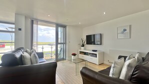 A modern living room with a glass coffee table, leather sofas, a white entertainment unit, and a large window offering a view of the outdoors. - A CONTEMPORARY BEACHSIDE APARTMENT ON HEADLAND ROAD FISTRAL 5 MINUTE FROM BEACH  (Cornwall)