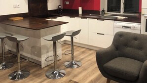 A modern kitchen with white cabinets, a wooden island, and bar stools. There is a grey armchair and a wooden coffee table with a remote control on it.