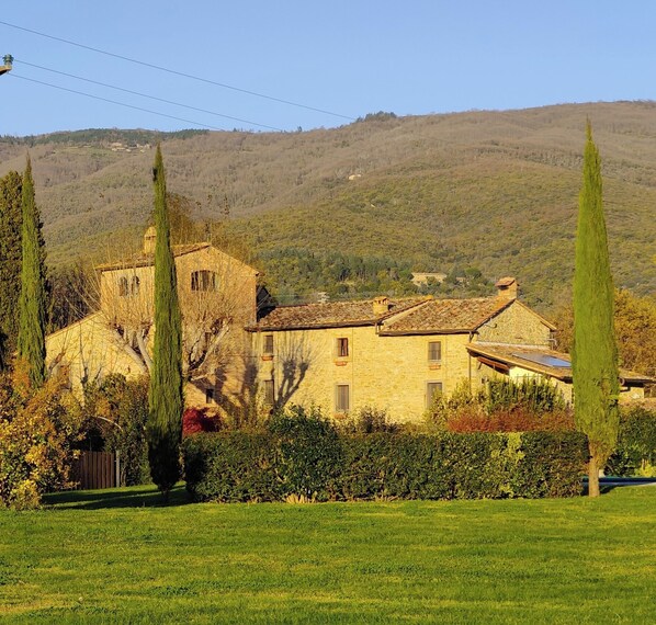 A stone building with a bell tower, surrounded by greenery and hills in the background.