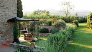 A stone building with a covered patio, a stone fireplace, and a garden with a lawn and various plants. - Large country apartment in Cortona 3bed3bath with shared pool & gorgeous gardens (Cortona)
