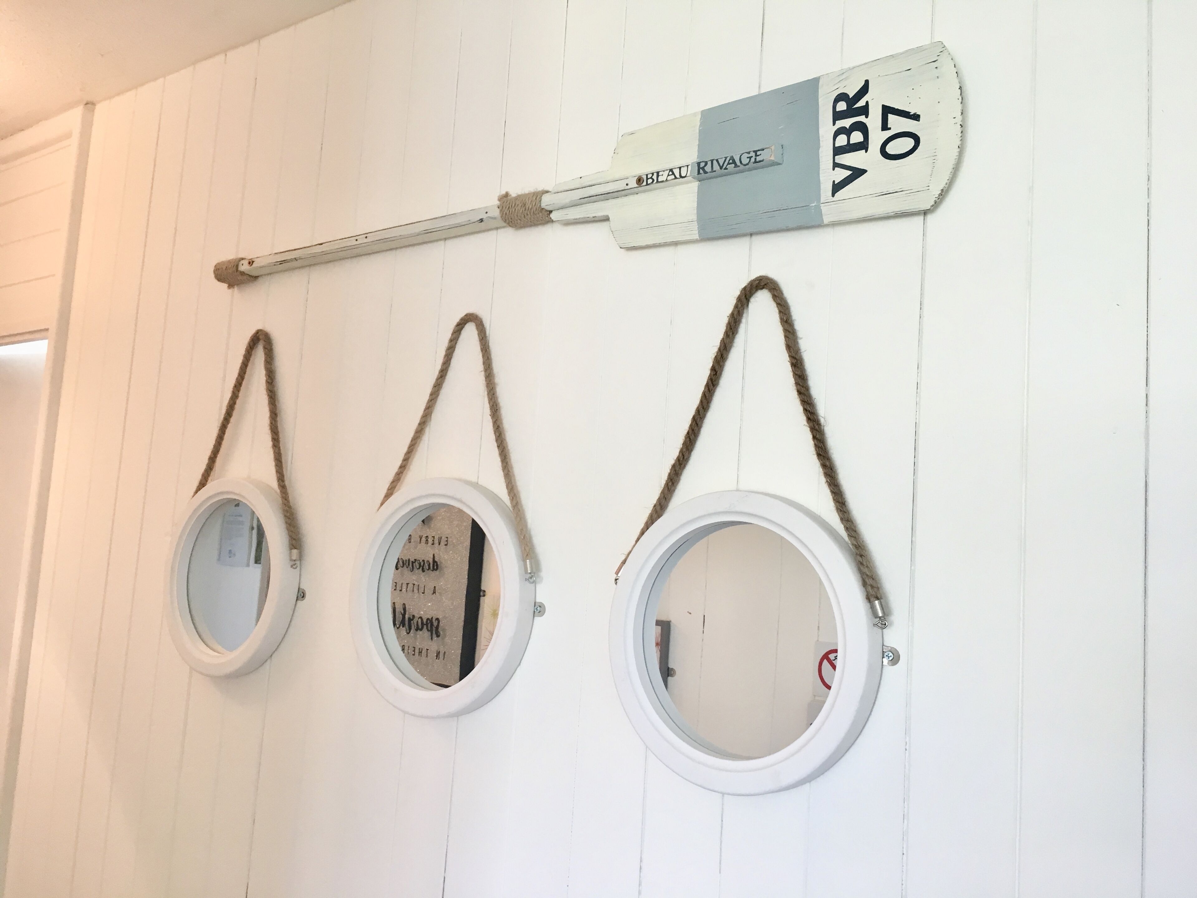 Trois miroirs ronds, suspendus à des cordes sur un mur blanc, une enseigne en bois au-dessus, et une porte sur la gauche.
