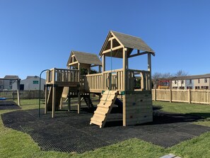 A wooden playground with climbing structures and a slide, surrounded by a fenced area and a grassy field.