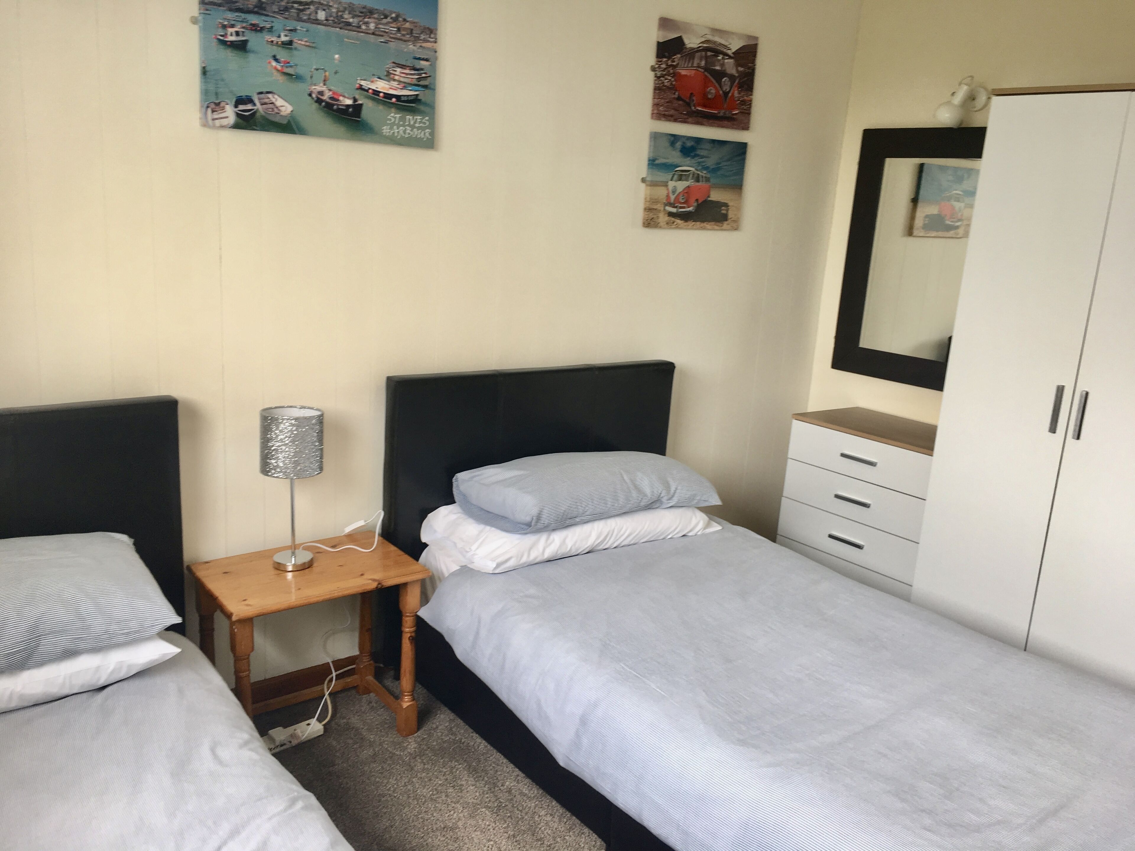 Een slaapkamer met twee enkele bedden, een nachtkastje met een lamp, een garderobe en twee aan de muur bevestigde foto's.