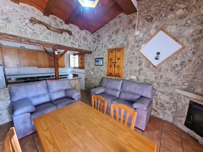 A cozy living area with a stone wall, a wooden ceiling, a kitchen in the background, and a fireplace.