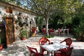 An outdoor patio with red plastic chairs and tables, surrounded by potted plants and a stone building with a rustic appearance.