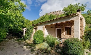 A rustic stone building with a tiled roof, surrounded by lush greenery and a clear sky.