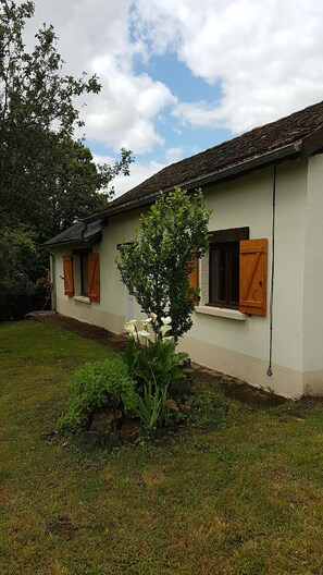 A single-story house with a tiled roof, a small garden with a tree and white flowers, and a clear sky. - Gite in quiet hamlet for rent. (Sarrazac)