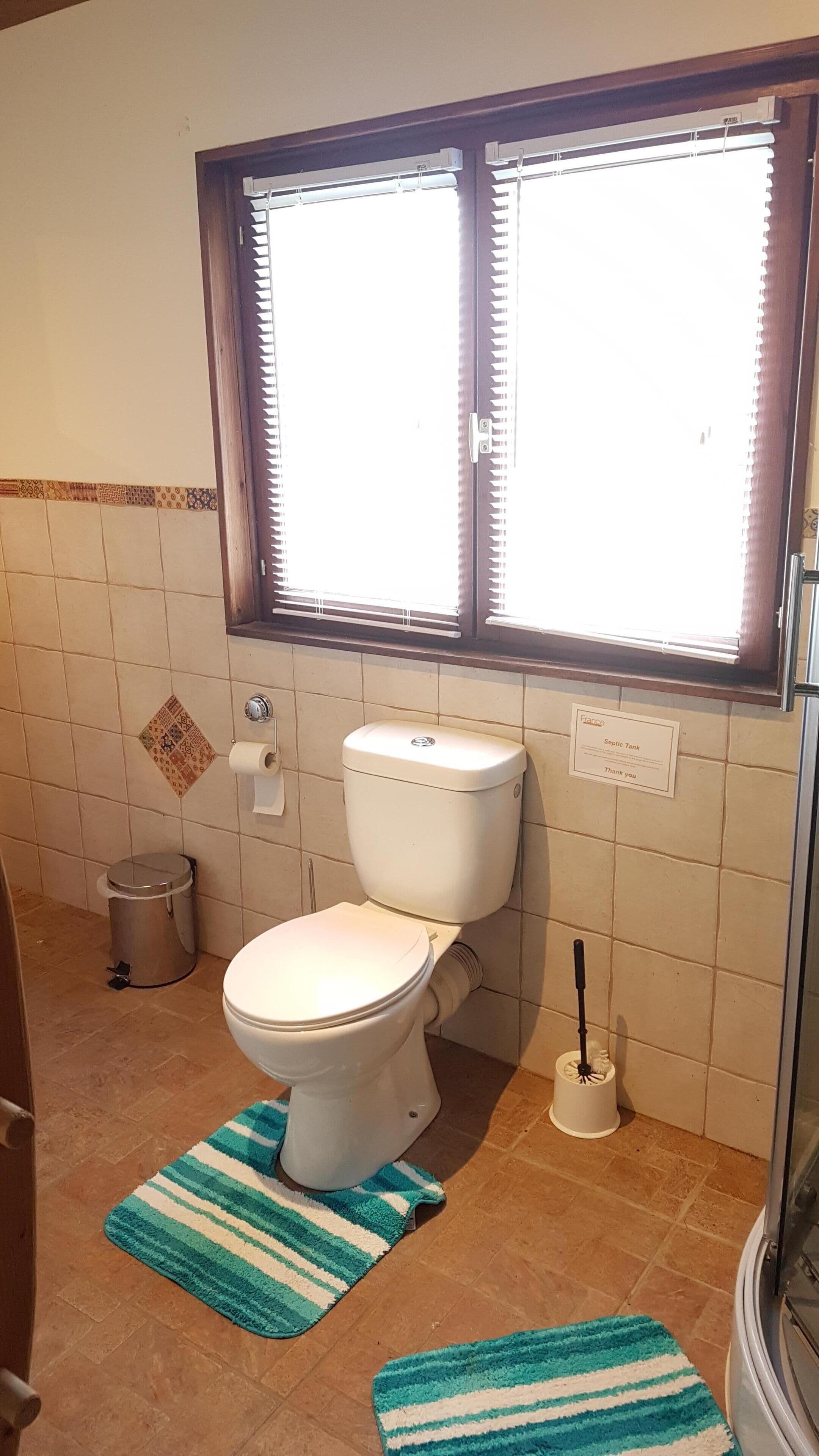 A bathroom with a toilet, a bidet, a trash can, and a window with blinds.