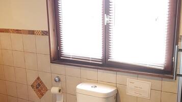 A bathroom with a toilet, a bidet, a trash can, and a window with blinds.