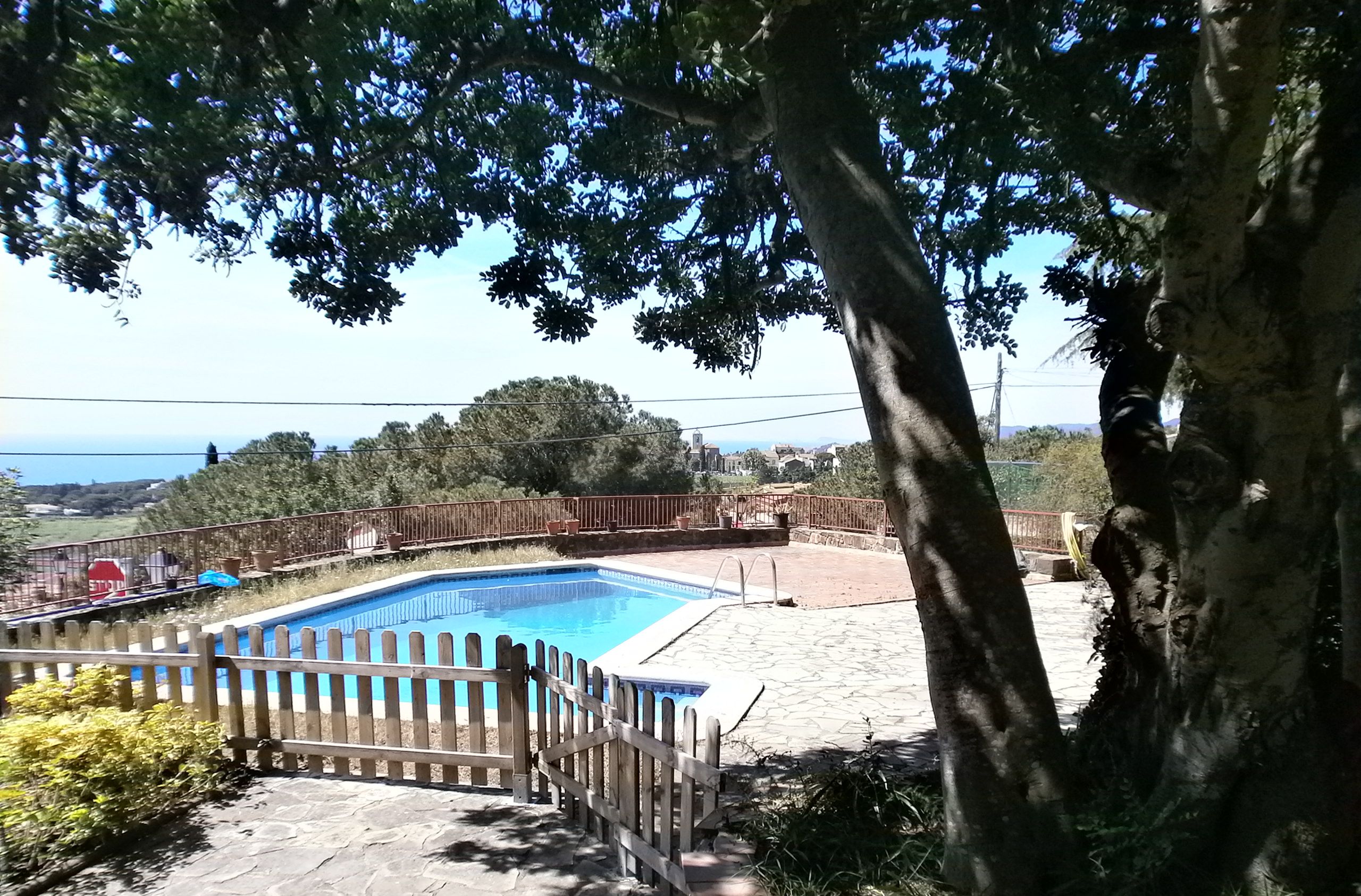 A swimming pool with a wooden fence, surrounded by trees and a view of the sea.
