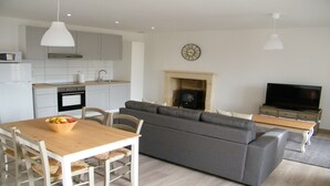 Eine moderne Küche mit weißen Schränken, einer Mikrowelle und einem Spülbecken. Ein Esstisch mit Stühlen und eine Schale Obst sind ebenfalls vorhanden. Im Wohnbereich befindet sich eine graue Couch und ein Fernseher.