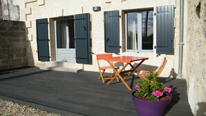 Eine Terrasse mit Holztisch und Stühlen, einem Blumentopf mit Blumen und einem Gebäude mit Fensterläden und einer Tür.