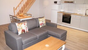Ein modernes Wohnzimmer mit einem grauen Ecksofa, einem Couchtisch aus Holz und einer Küchenzeile mit weißen Schränken und Geräten.