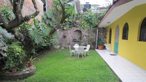 A small, enclosed garden with a stone wall, a yellow building with arched windows, and a white outdoor seating area.