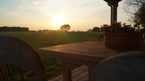 A wooden table and chairs under a covered area with a view of a sunset over a grassy field. - Holiday home 't Ligt ter velde (Lichtervelde)