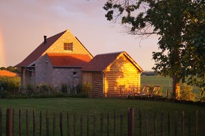 Een landelijke schuur met een bakstenen uitbouw en een houten tuinhuisje, omgeven door een omheind erf en een boom.