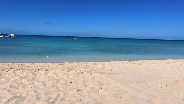 Une plage avec une eau d’un bleu limpide, un rivage sablonneux et quelques bateaux à l’horizon.