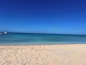 A beach with clear blue water, a sandy shore, and a few boats in the distance.