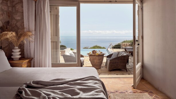 A bedroom with a large bed, a view of the sea through an open door, a wicker chair, and a table with breakfast items.