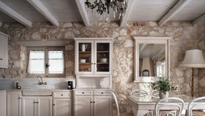 A rustic kitchen with stone walls, white cabinetry, a marble countertop, and a dining area with white chairs and a table.