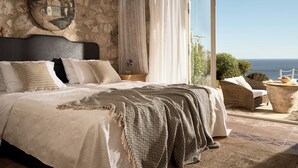 A bedroom with a stone wall, a bed with multiple pillows, a nightstand, and a view of the sea.