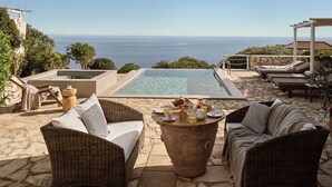 A poolside area with wicker furniture, a stone table with drinks, and a view of the sea.