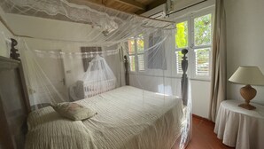 Ein Schlafzimmer mit Baldachinbett, Nachttisch mit Lampe und Blick durch ein Fenster mit Rollläden.