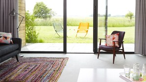 Una sala de estar moderna con un sofá, una alfombra colorida y un ventanal de vidrio que da a un área exterior con césped y dos sillas.