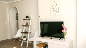 Un salon doté d’un téléviseur à écran plat, de chaises blanches, d’une console blanche et d’une étagère murale agrémentée d’objets décoratifs.