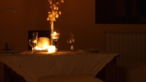 Un comedor tenuemente iluminado con una mesa dispuesta para dos personas, que incluye una vela, un florero con flores secas y dos copas de vino.