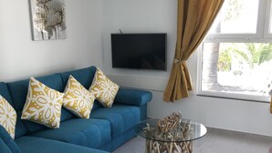 Un salon avec un canapé bleu, des coussins à motifs, un téléviseur à écran plat, une table basse en verre et une fenêtre avec des rideaux.