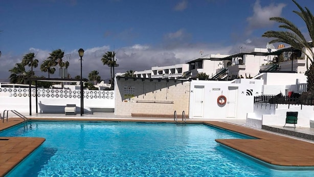 Une piscine d’un bleu éclatant, avec une terrasse en bois, des murs blancs et des palmiers en arrière-plan.