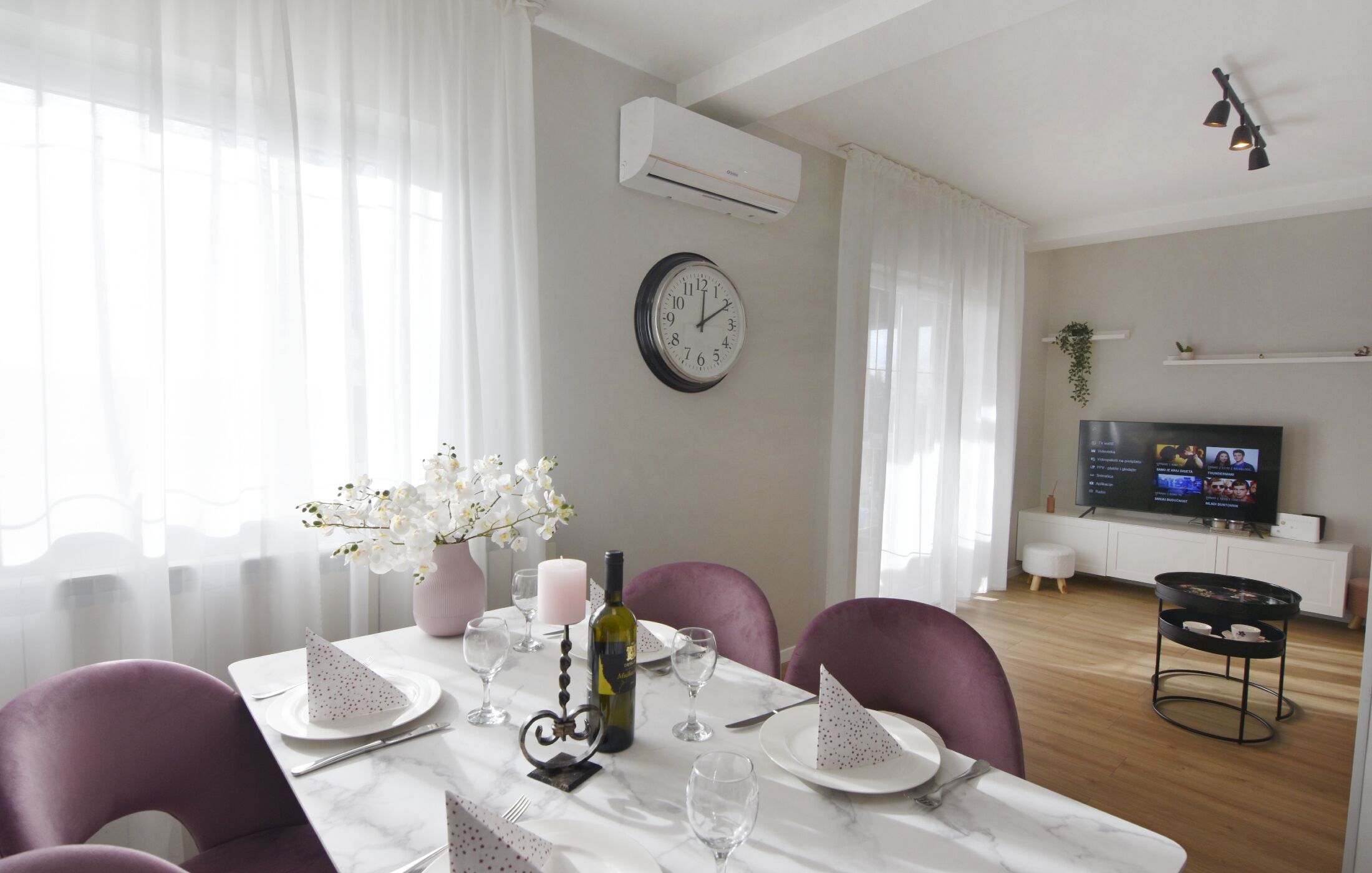 A dining area with a white table set for two, a clock on the wall, and a TV in the background.