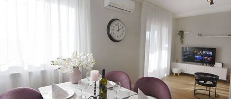A dining area with a white table set for two, a clock on the wall, and a TV in the background.