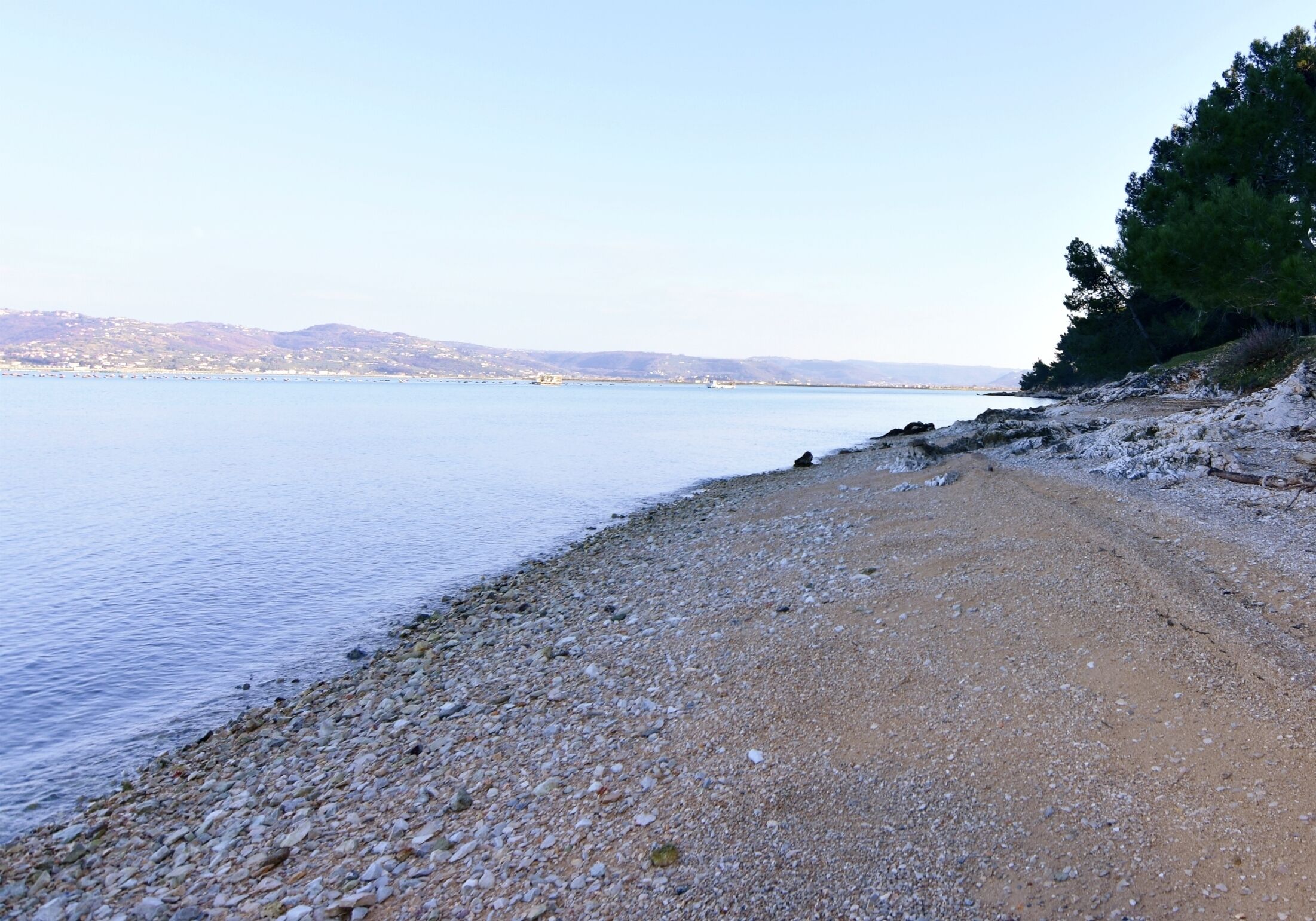 A pebbly beach with calm water and distant hills.