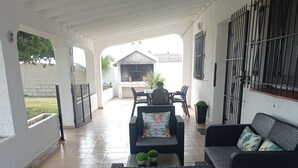 Un espace patio extérieur couvert, aménagé avec un coin salon comprenant un canapé et deux fauteuils, une petite table avec une plante, et un barbecue à l’arrière-plan.