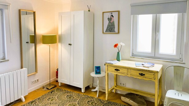 Una habitación con un armario blanco, un escritorio con cajones, un taburete pequeño, un radiador y una alfombra estampada.