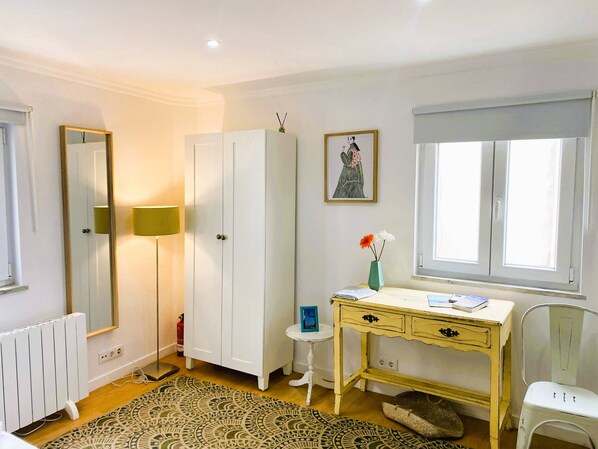Een kamer met een witte kast, een bureau met lades, een klein krukje, een radiator en een kamerkleed met dessin.
