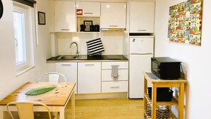 A compact kitchen with white cabinets, a stainless steel sink, a black and white striped towel, a refrigerator, and a wooden table with a green leaf decoration.