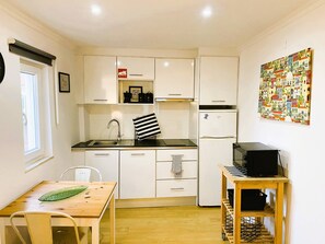 Una cocina compacta con armarios blancos, un lavamanos de acero inoxidable, una toalla a rayas negras y blancas, un refrigerador y una mesa de madera con una decoración de hoja verde.