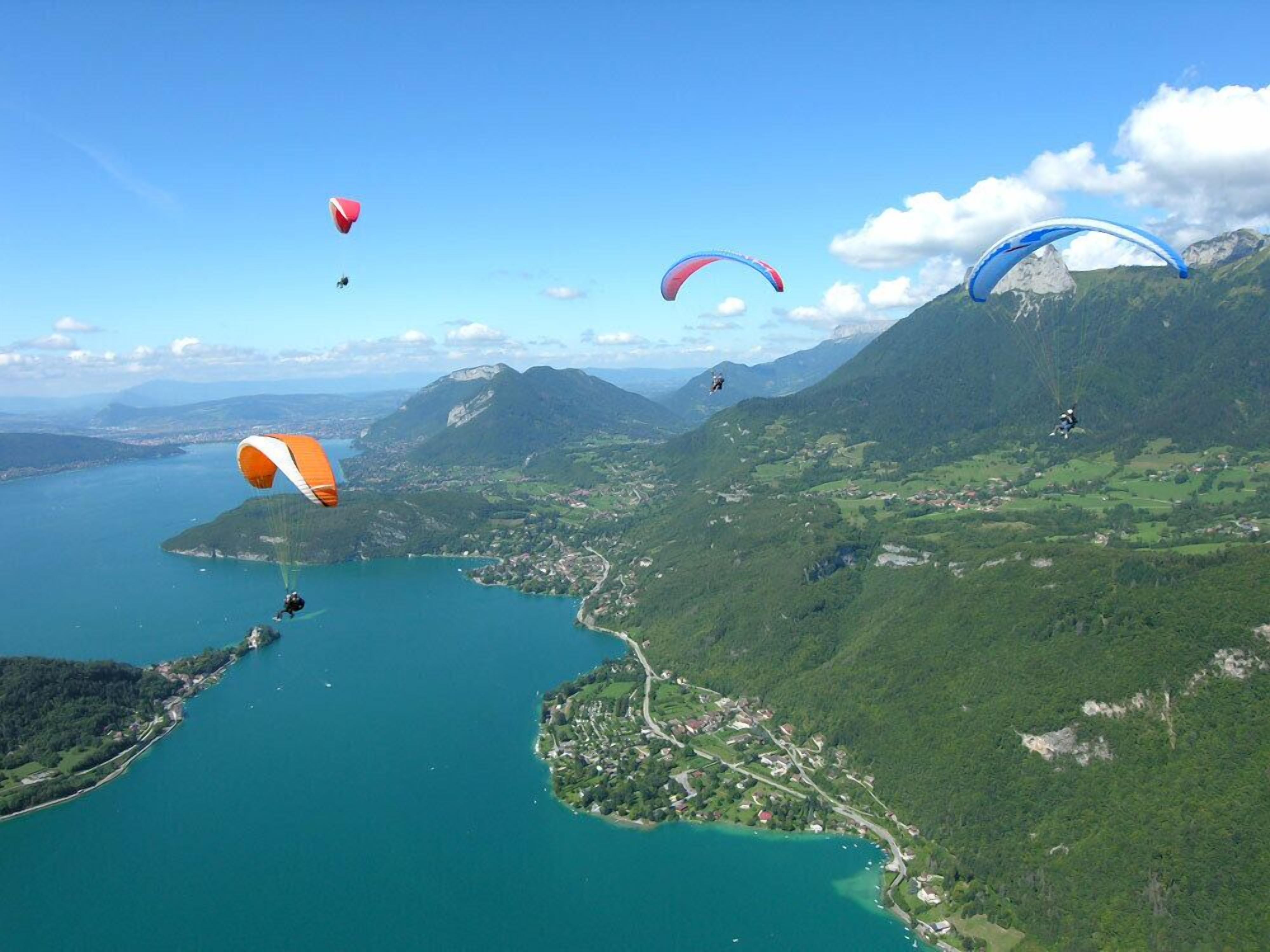 Eine Küstenlandschaft mit einem See, Bergen und mehreren Gleitschirmfliegern am Himmel.
