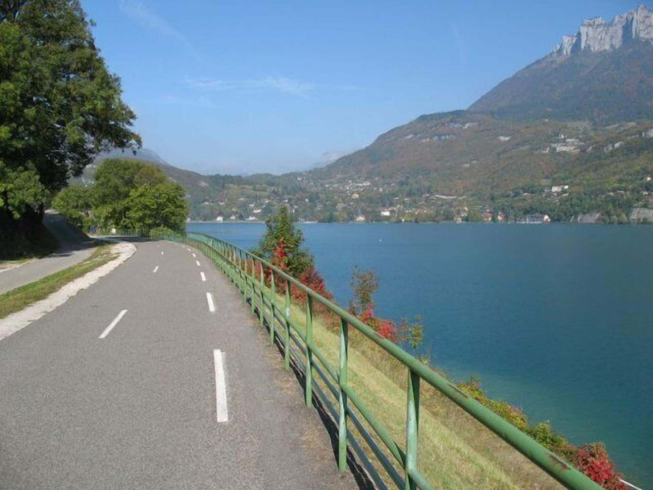 Una carretera con barandilla verde, un lago y una montaña al fondo.
