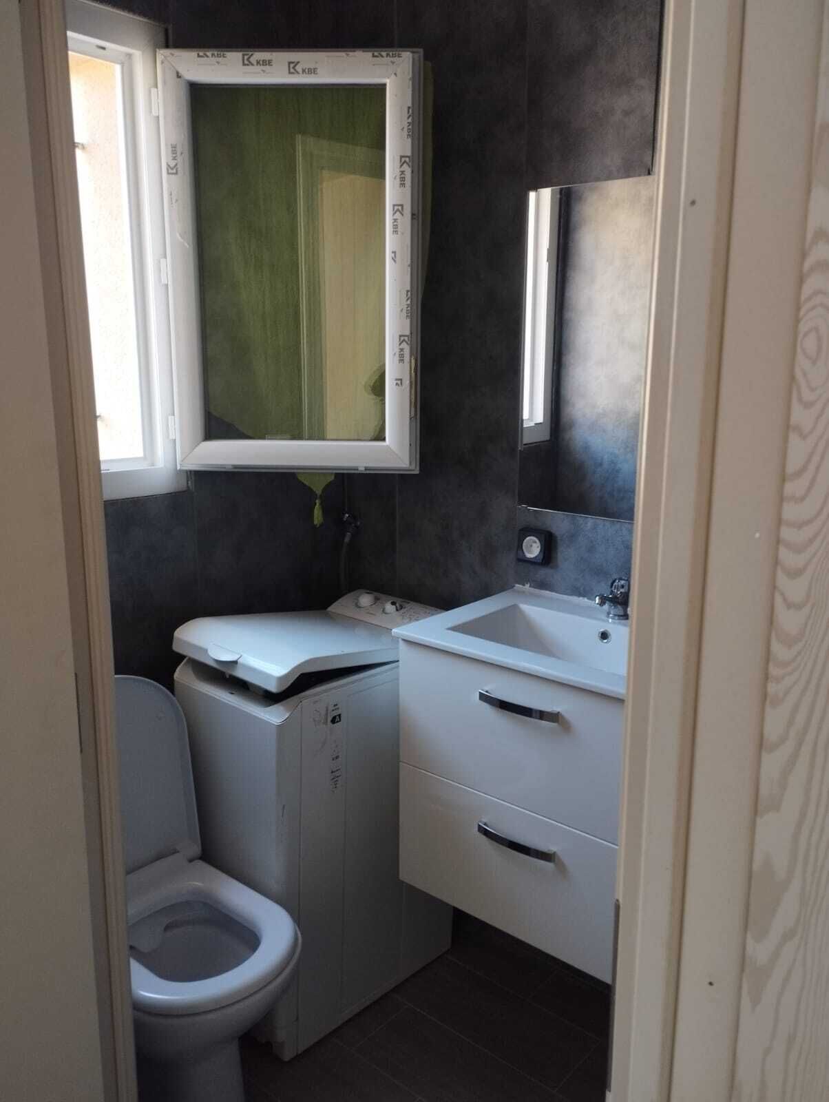 A compact bathroom with a toilet, a washing machine, a sink, and a mirror.