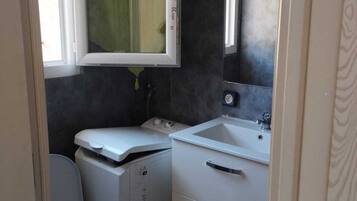 A compact bathroom with a toilet, a washing machine, a sink, and a mirror.