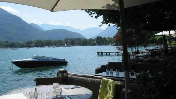 An outdoor dining area with a view of a lake and mountains. There are tables and chairs under umbrellas, and a boat is visible on the water.