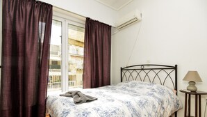 Um quarto com cama, janela com cortinas, mesa de cabeceira com abajur e ar condicionado na parede.
