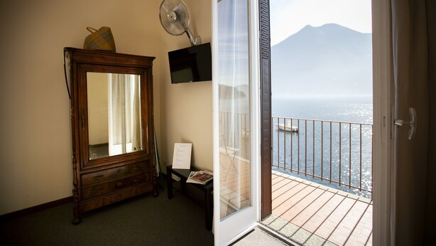 A room with a wooden dresser, a mirror, a fan, and a balcony with a view of the sea and a mountain. - La Darsena di Riva Grande - Comballi (montrasio)