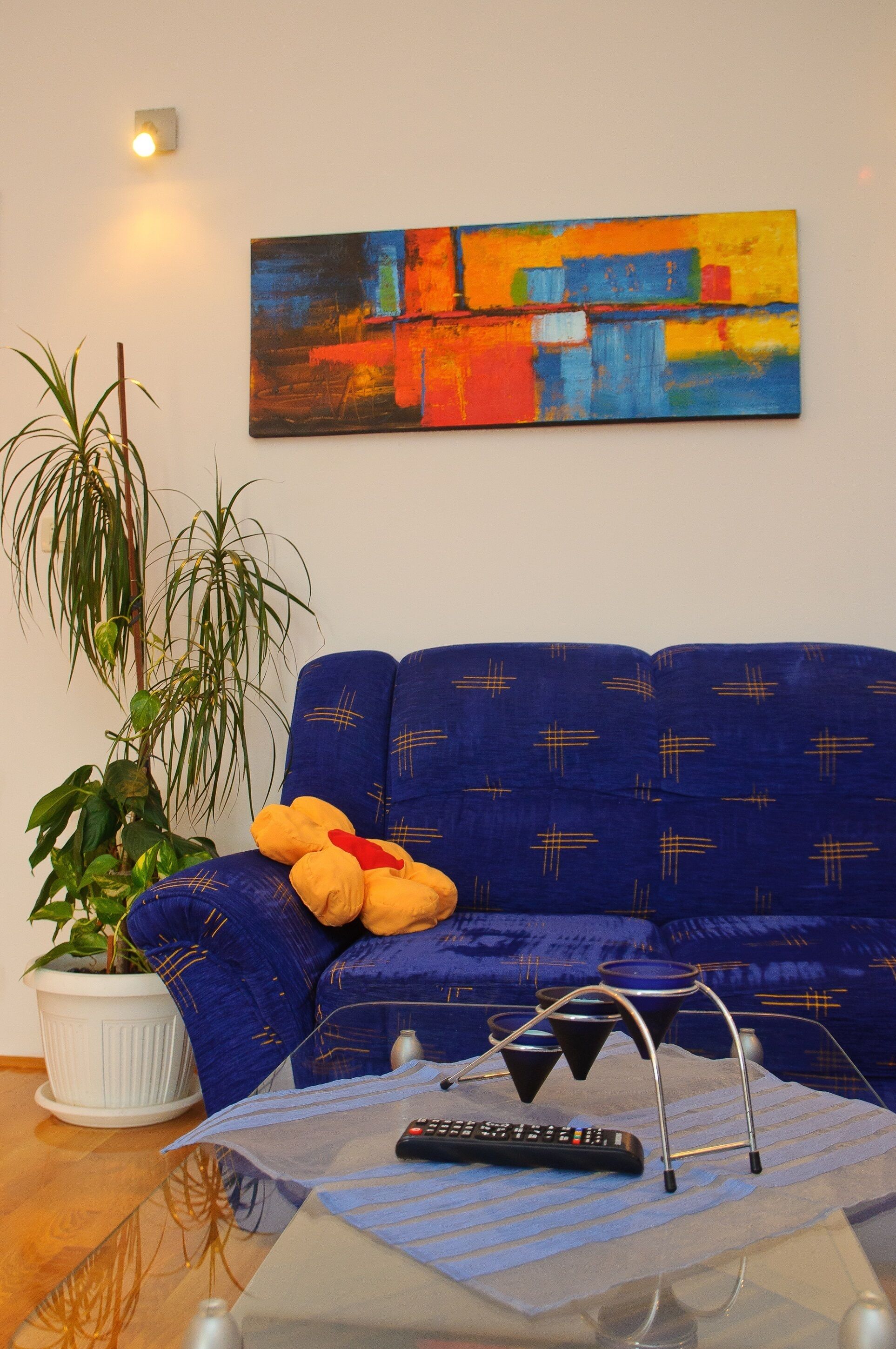 Un salon avec un canapé bleu, une plante en pot, une télécommande et un tableau abstrait aux couleurs vives accroché au mur.