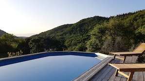 An infinity pool with wooden deck chairs, surrounded by lush greenery and hills.