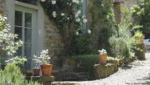 A stone building with a climbing white rose bush, potted plants, and a gravel driveway.